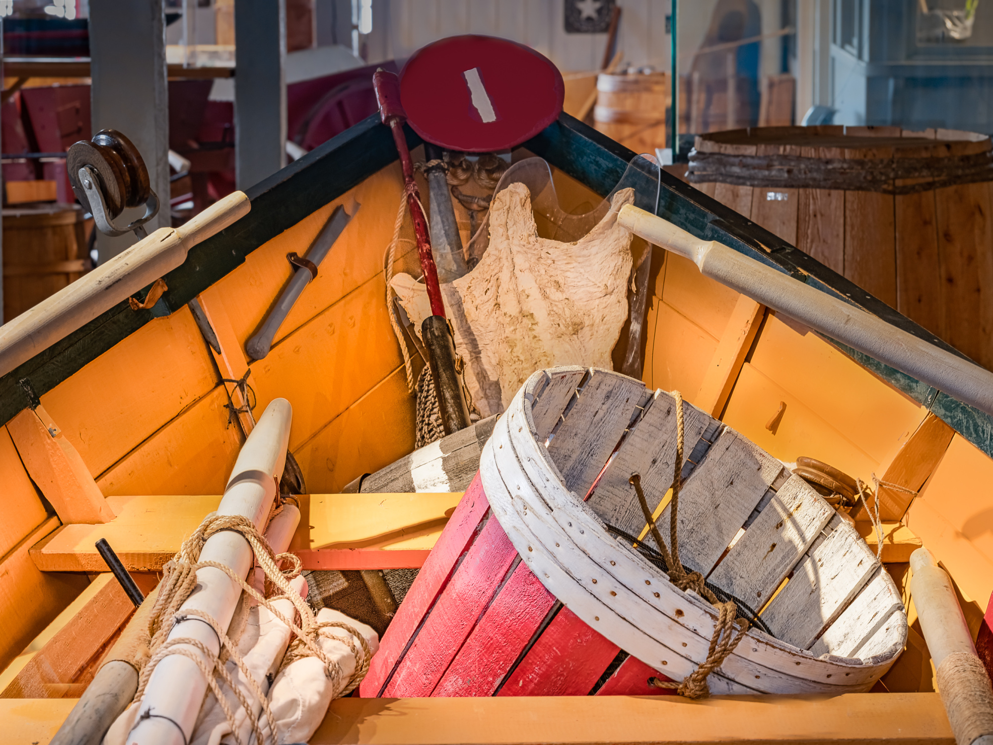 A close-up view looking into the bow of a traditional wooden fishing dory with yellow-painted interior boards and green trim. The boat is packed with historic fishing gear, including a red-and-white wooden trawl tub, bundled canvas sails tied with rope, wooden oars rested along the sides, and red marker buoys.