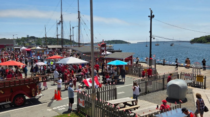 An elevated photograph of a vibrant Canada Day celebration at a historic waterfront. A large crowd dressed in red fills a wharf area next to two multi-masted tall ships. A prominent Canadian flag waves at the top left, with other flags throughout the scene. Picnic tables, a red fire engine, and a bay with sailboats under a blue sky complete the view.