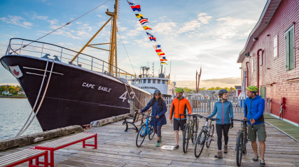 A group of four cyclists walk their bikes along a wooden boardwalk at sunset, passing the large black-hulled ship "Cape Sable" and a bright red building.