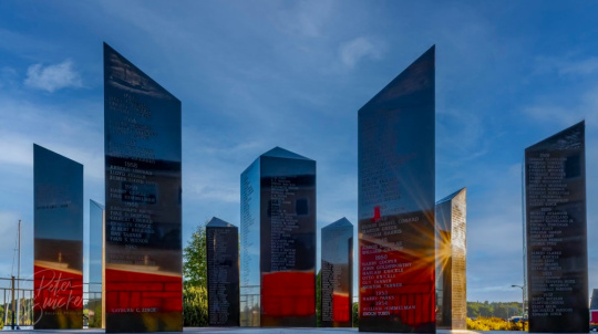 Fisher's memorial, a circle of black granite memorial pillars, engraved with names and dates, stands on a stone base with a compass rose design overlooking a peaceful harbor.